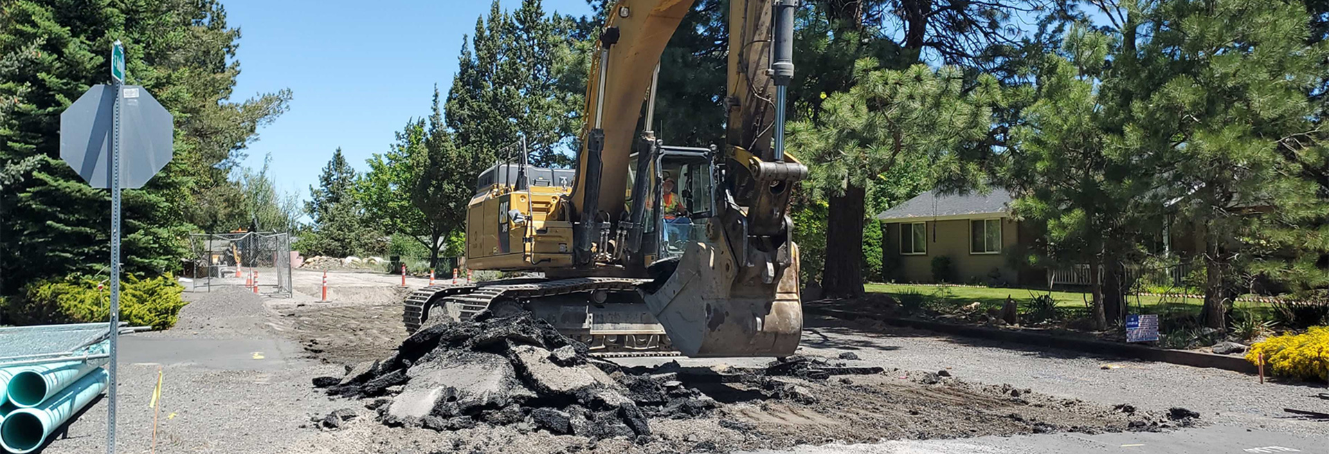 Yellow excavator digging into a city street. Houses and trees surround the street.