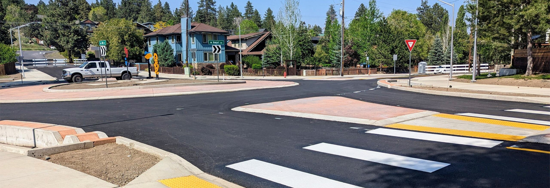 Roundabout shown from street level. Black asphalt, gray sidewalks, street signs, and houses surrounding.