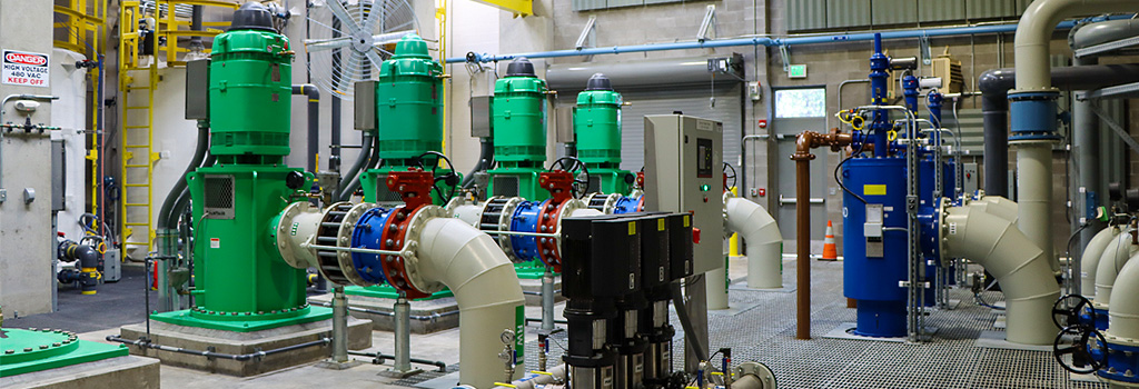 Inside the water filtration facility, green pump heads and white pipes inside a gray block structure.