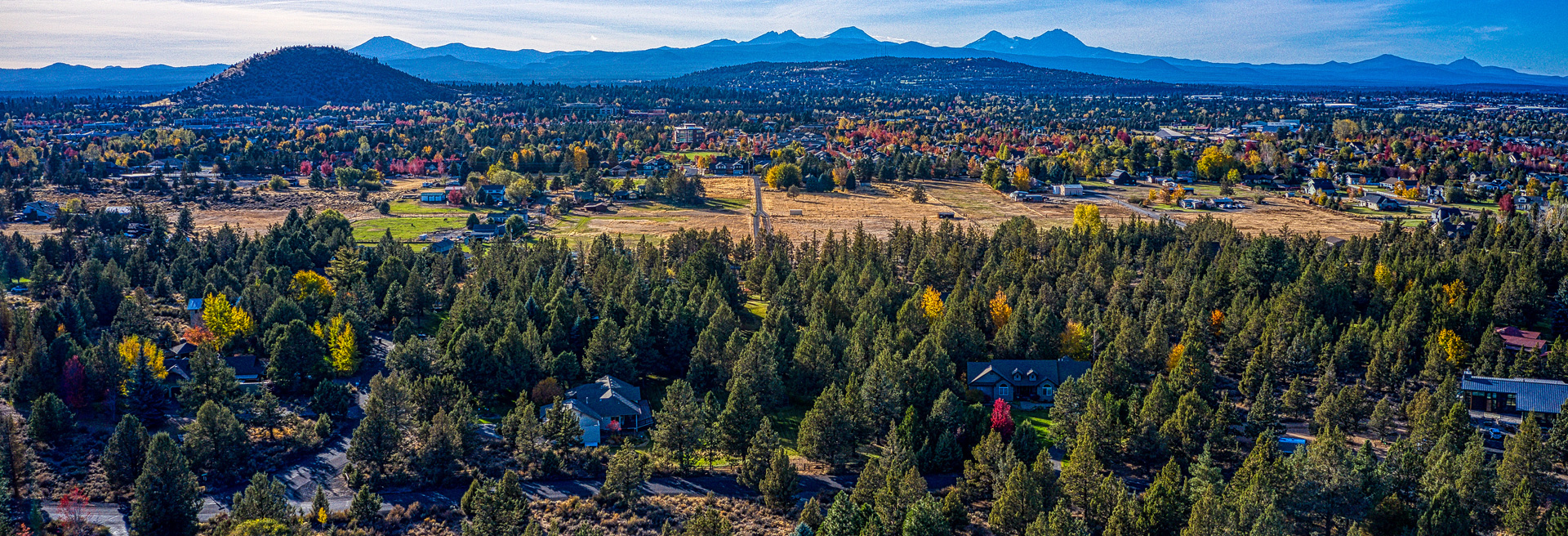 Drone shot of Bend looking west. Pilot Butte in the mid-ground. Party cloudy skies. Trees, roads, and houses.