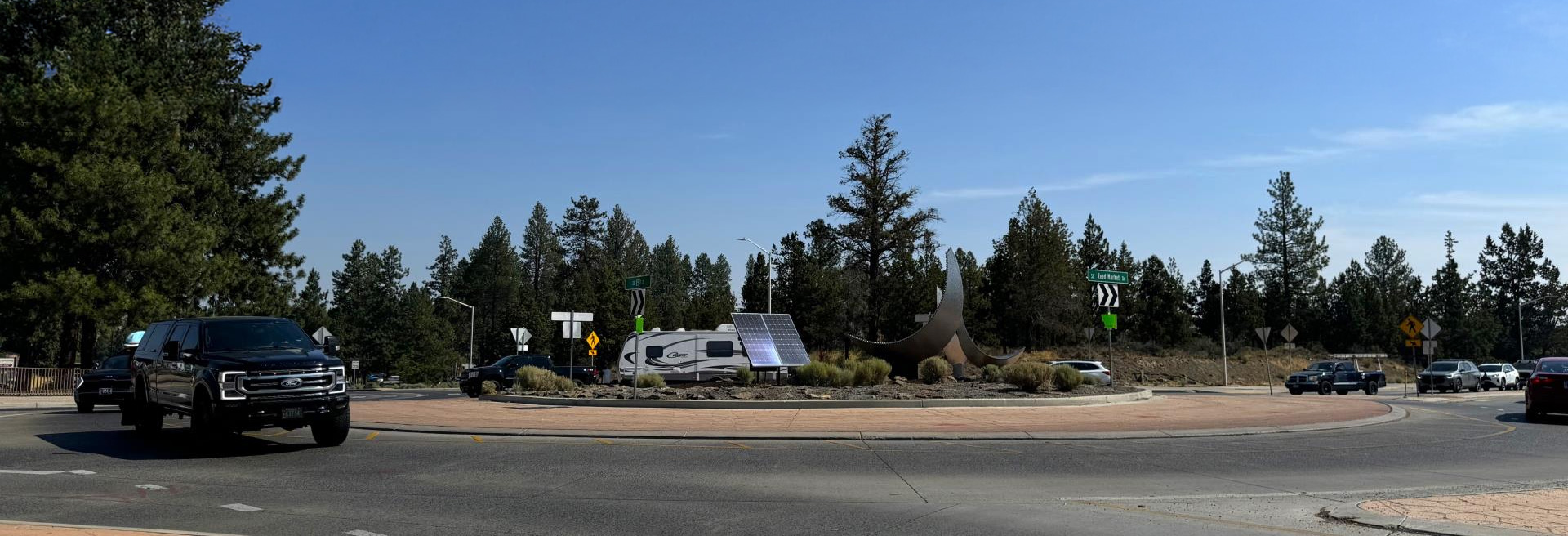 Street view of Traffic circle, cars and trucks entering and exiting the circle. Trees and blue sky in the background.