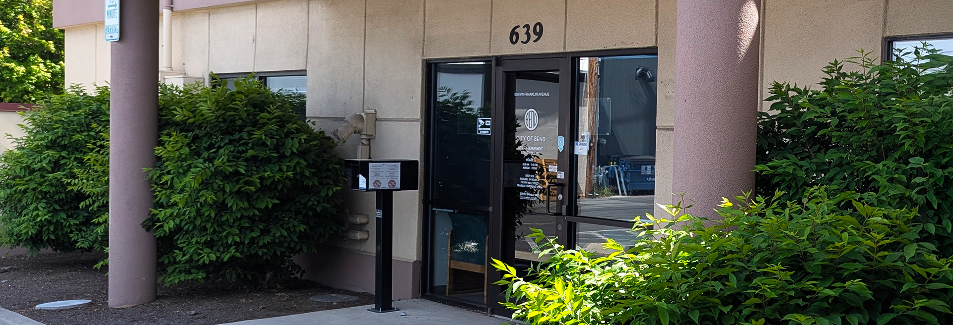 Main Entrance to Water Services Billing building. Tan siding, green plants in front by the glass entry door.