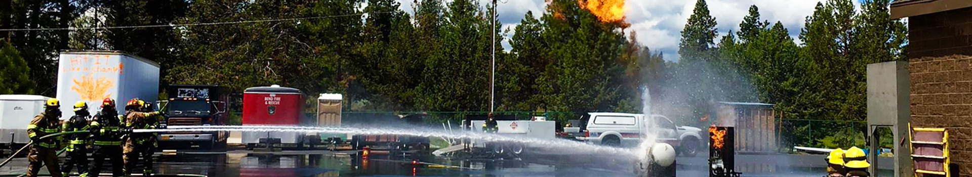 Firefighters spraying water onto a propane tank that is emitting fire from it.